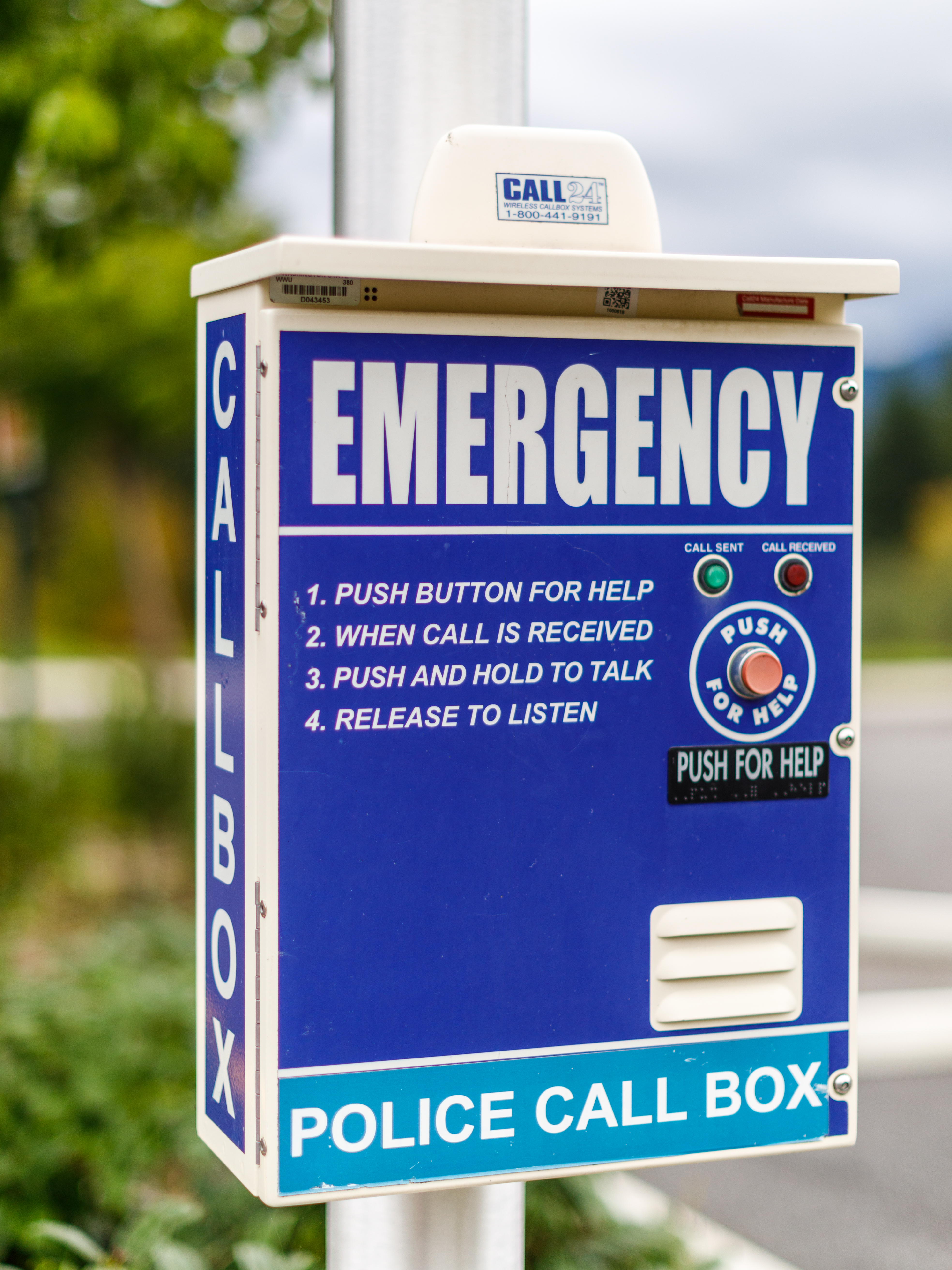 Photograph of Western’s blue 24 hour emergency police call box. Box reads in large letters, emergency, call box and police call box. Instructions for its use say “1. Push button for help 2. When call is received 3. Push and hold to talk 4. Release to listen” On the right side of the box there is a large red button, labeled with “Push for Help” with the braille underneath.