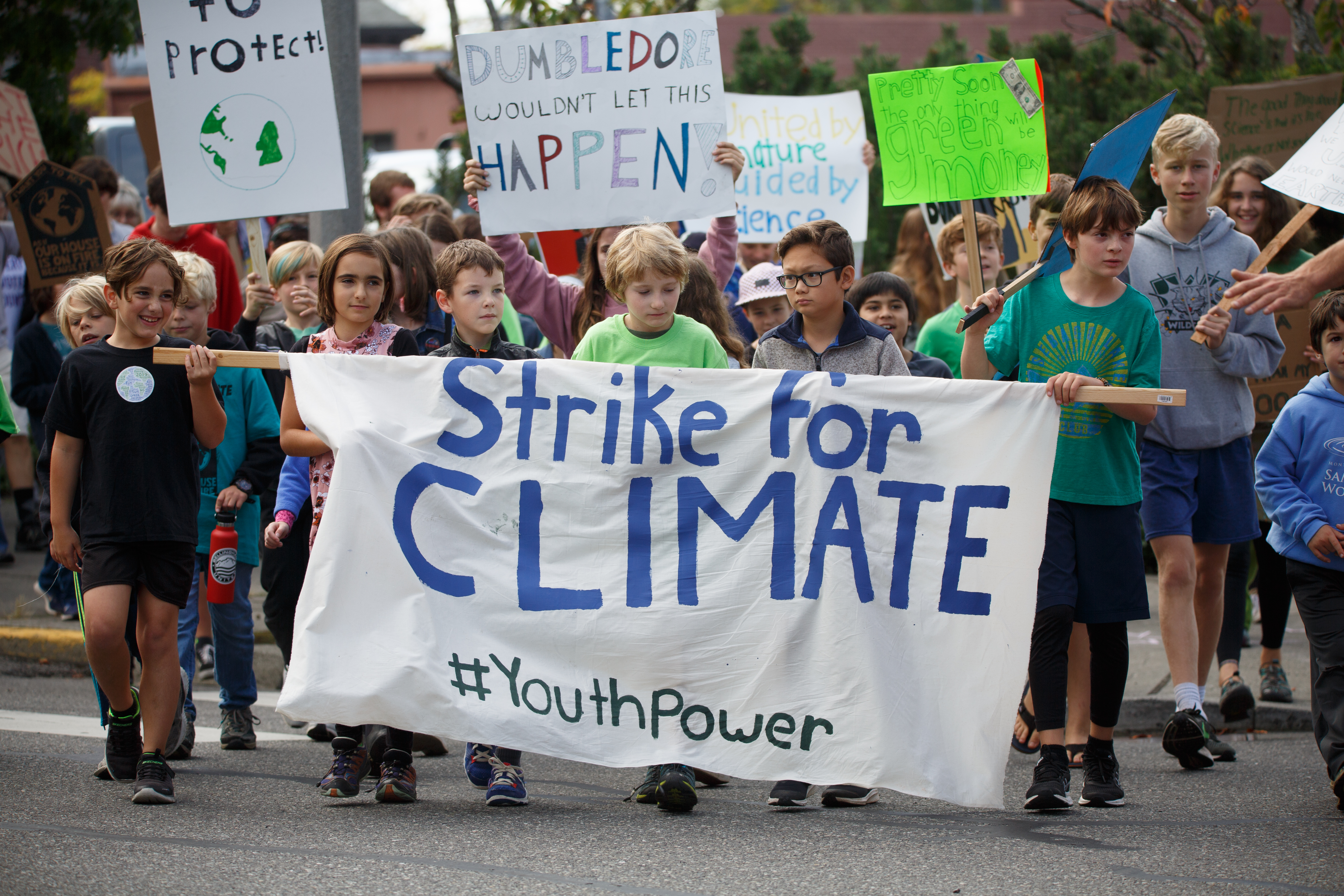 Image of six young students approximately elementary age carry a white banner that reads “Strike for Climate #youthpower.” Signs are held up behind the students, two of which can be read completely, “Dumbledore wouldn’t let this happen” and “Pretty soon the only green thing will be money.”