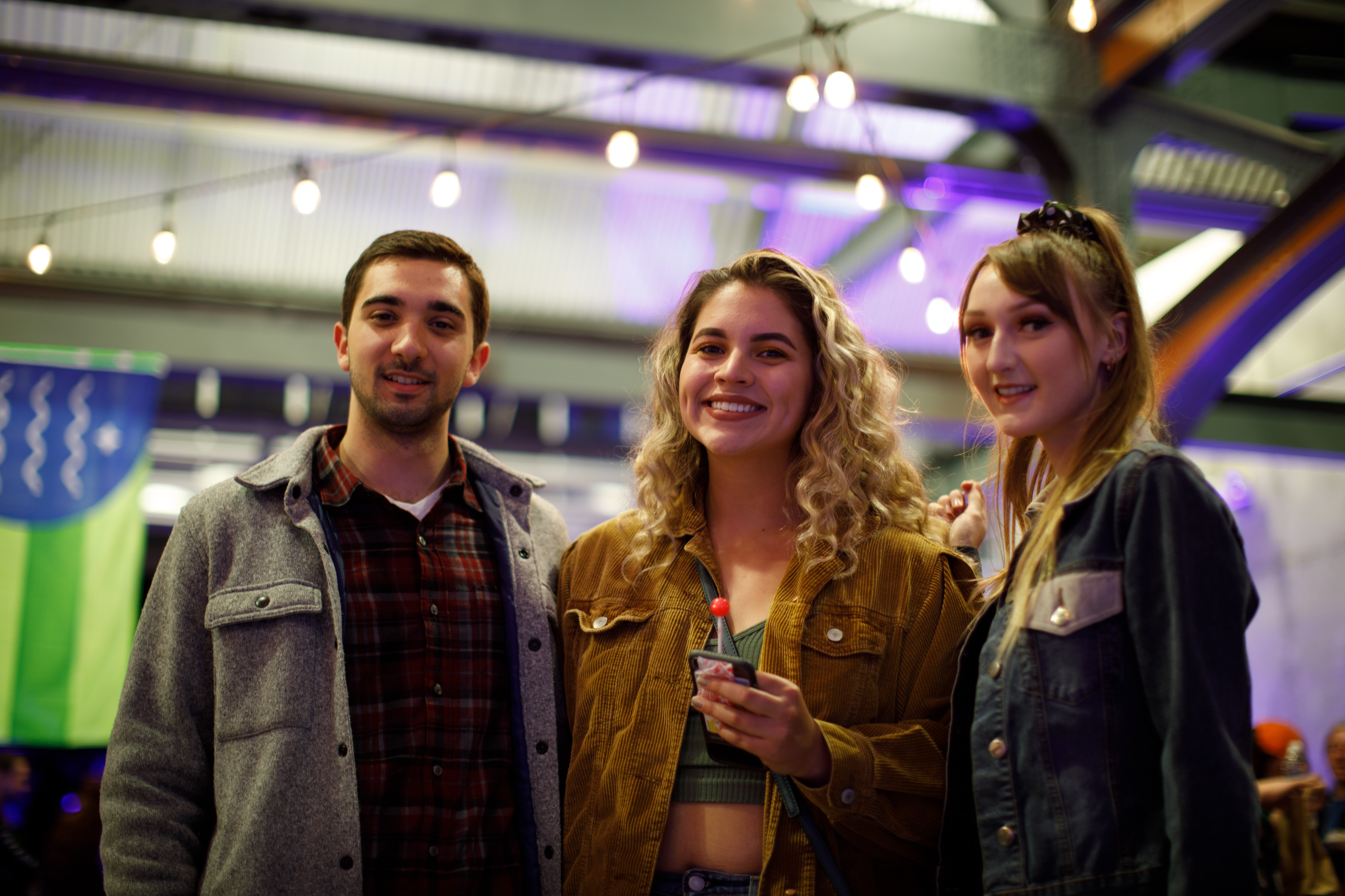 Image of three students standing shoulder to shoulder and smiling together, the indoor space of the Market Depot Square can be seen in the background, including a hanging Bellingham city flag.