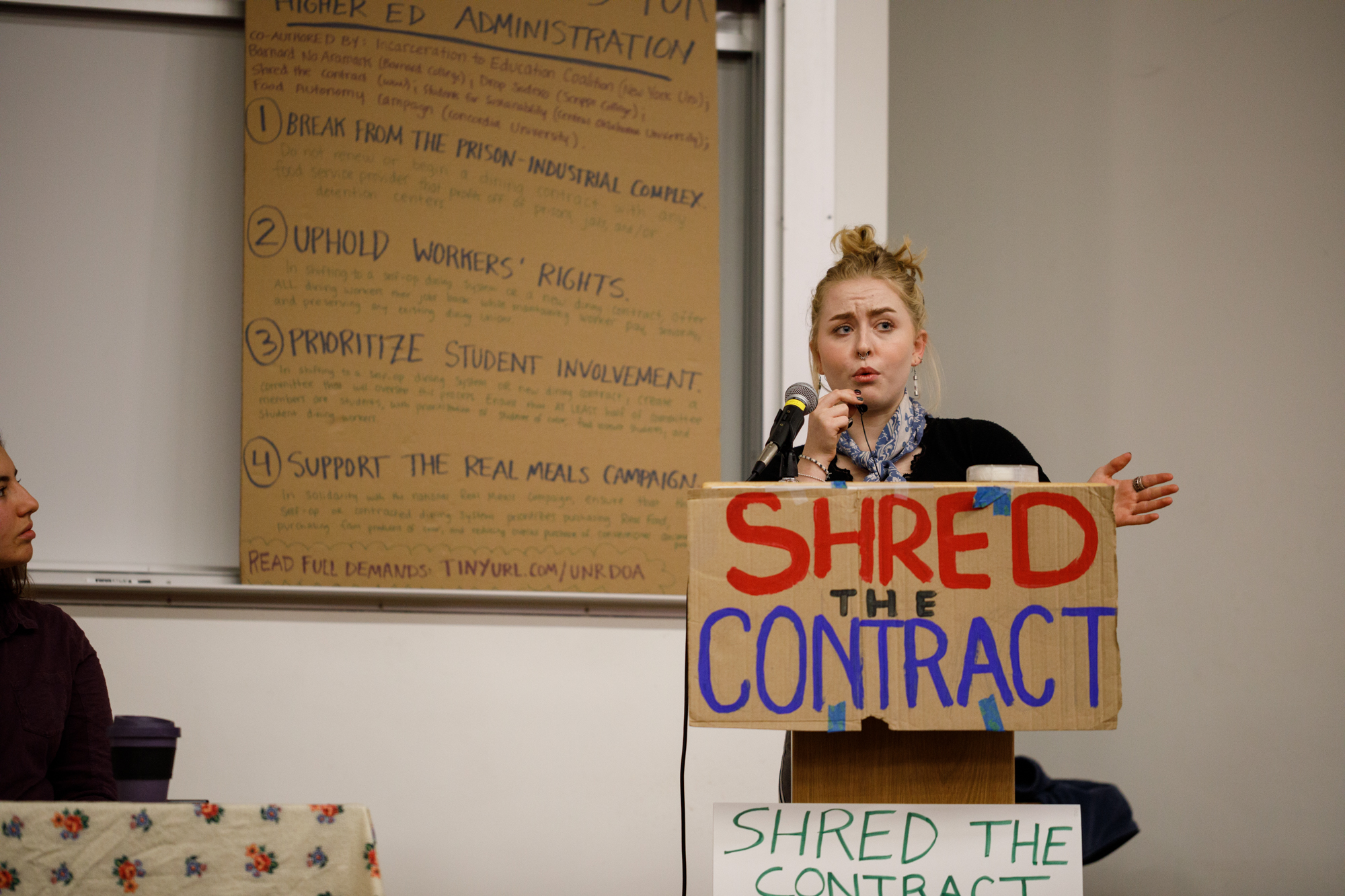 Image of Sophia Gamble speaking from the podium. The sign on the podium reads “Shred the Contract”.