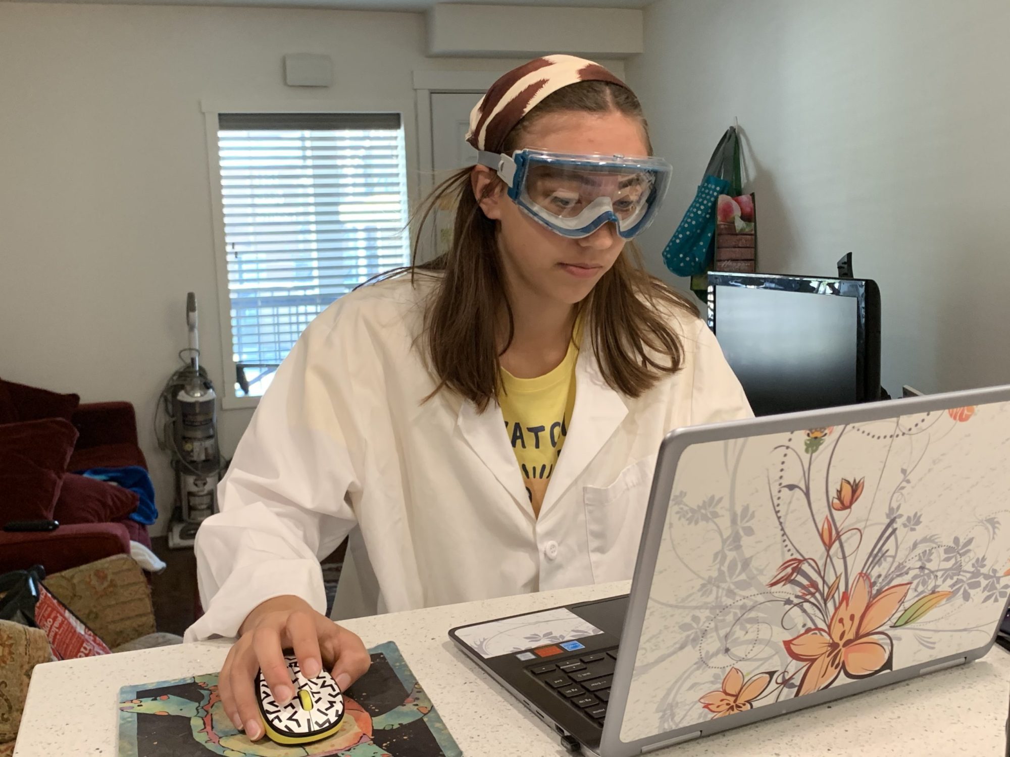 Image of a female student sitting in front of a computer in a lab coat and goggles.